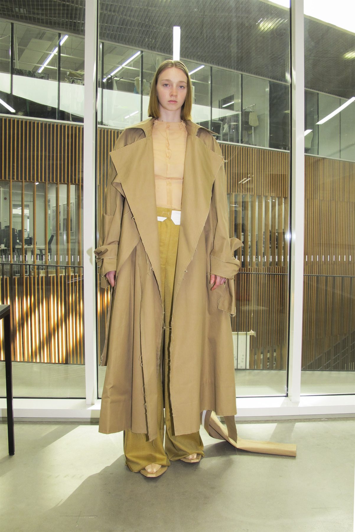 Model wearing a long trenchcoat with sheer top and sand-colored trousers