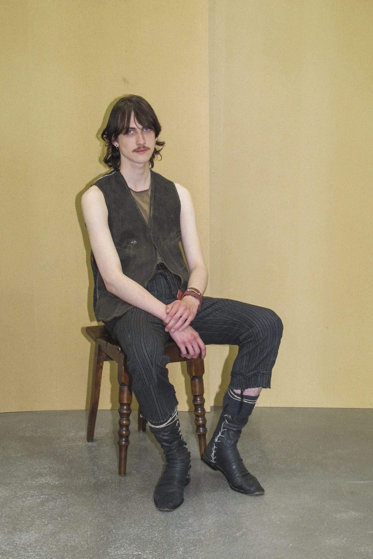 Photo of model sitting and wearing pinstripe trousers, khaki vest and sheer black shirt