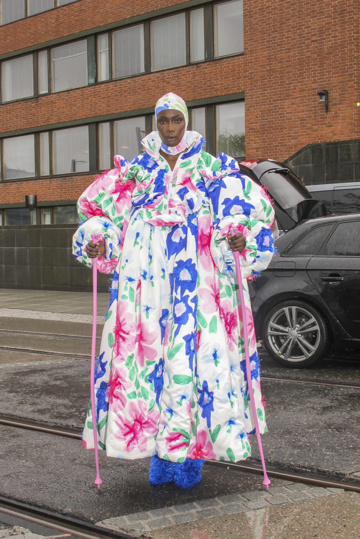 Model wearing a huge puffy dress with puffy sleeves and extensive flower print. Pink alpine skiing poles as accessories