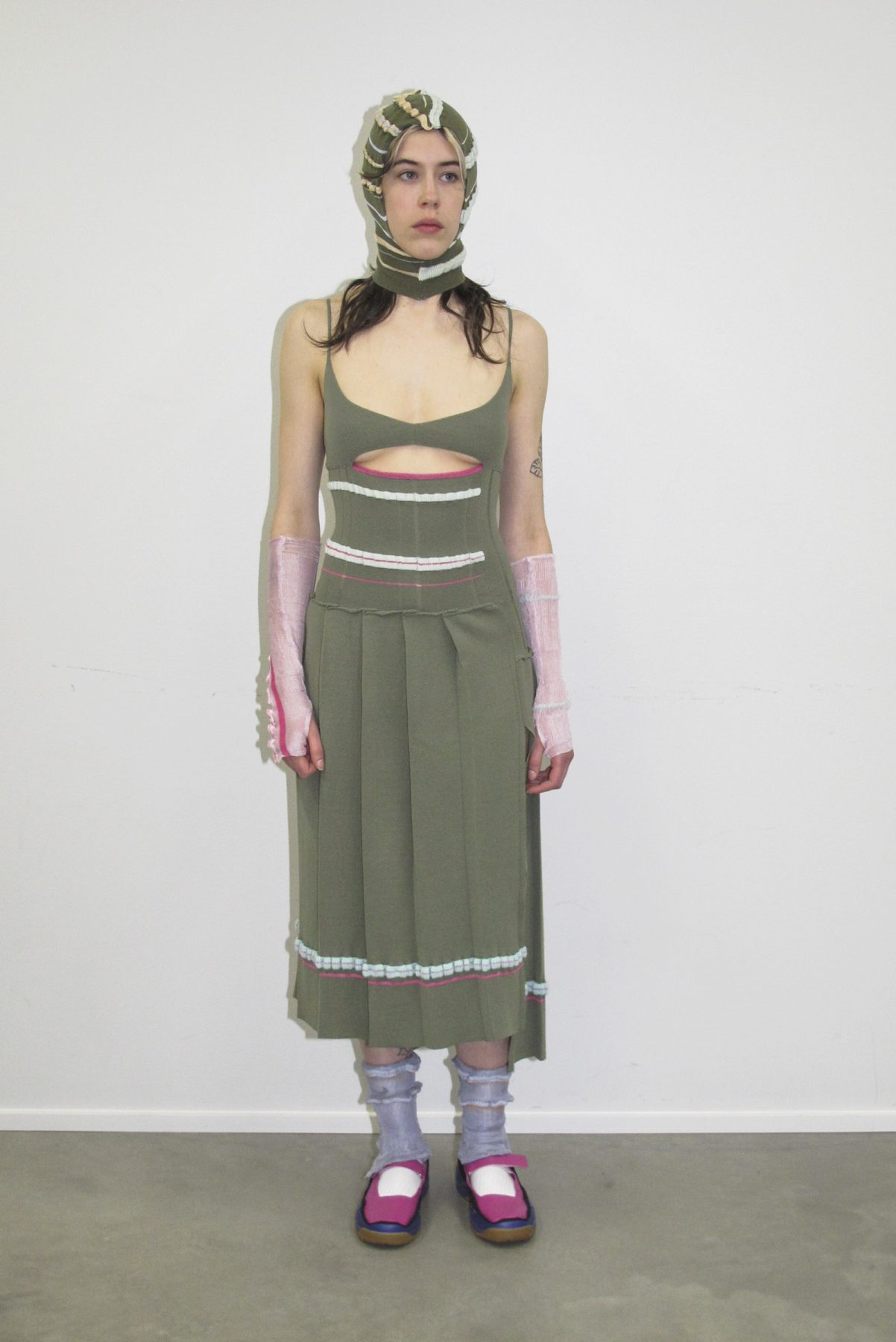 Model wearing a green knitted striped dress, matching balaclava, lilac socs and pink shoes