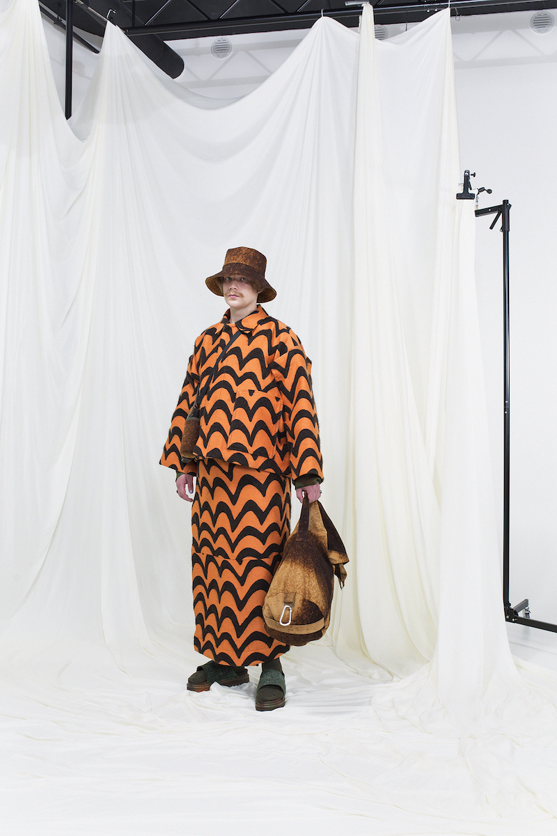 Model wearing a born & orange jacquard jacket with matching skirt, brown rug sack and a matching bucket hat.
