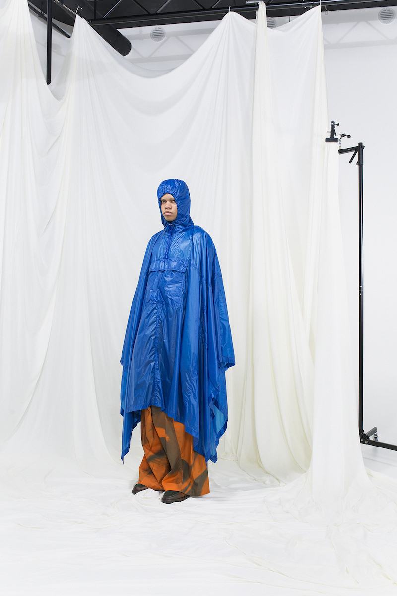 Model wearing a hood blue nylon raincoat with orange printed trousers underneath.
