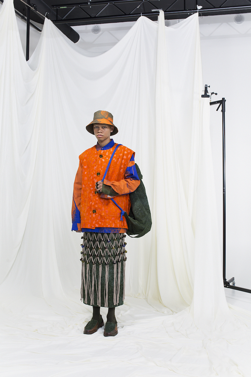 Model wearing a orange coat, geen skirt with macrame overskirt, green rug sack and a bucket hat