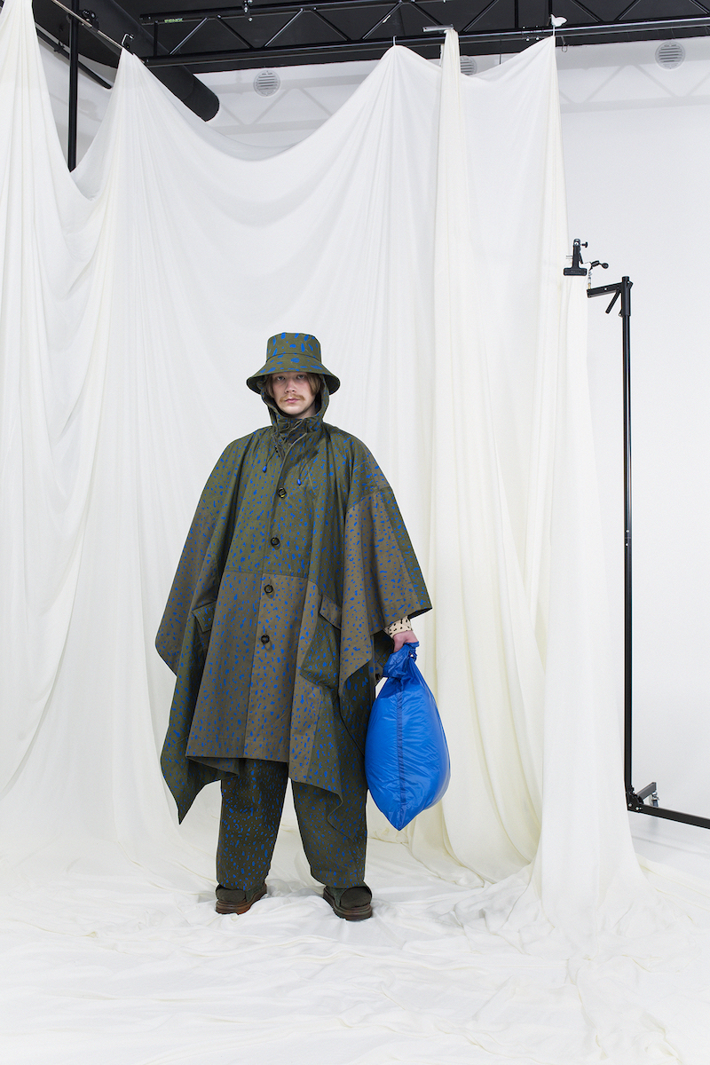 Model wearing a green hooded raincoat with blue dotted print, matching trousers, blue nylon bag and a bucket hat