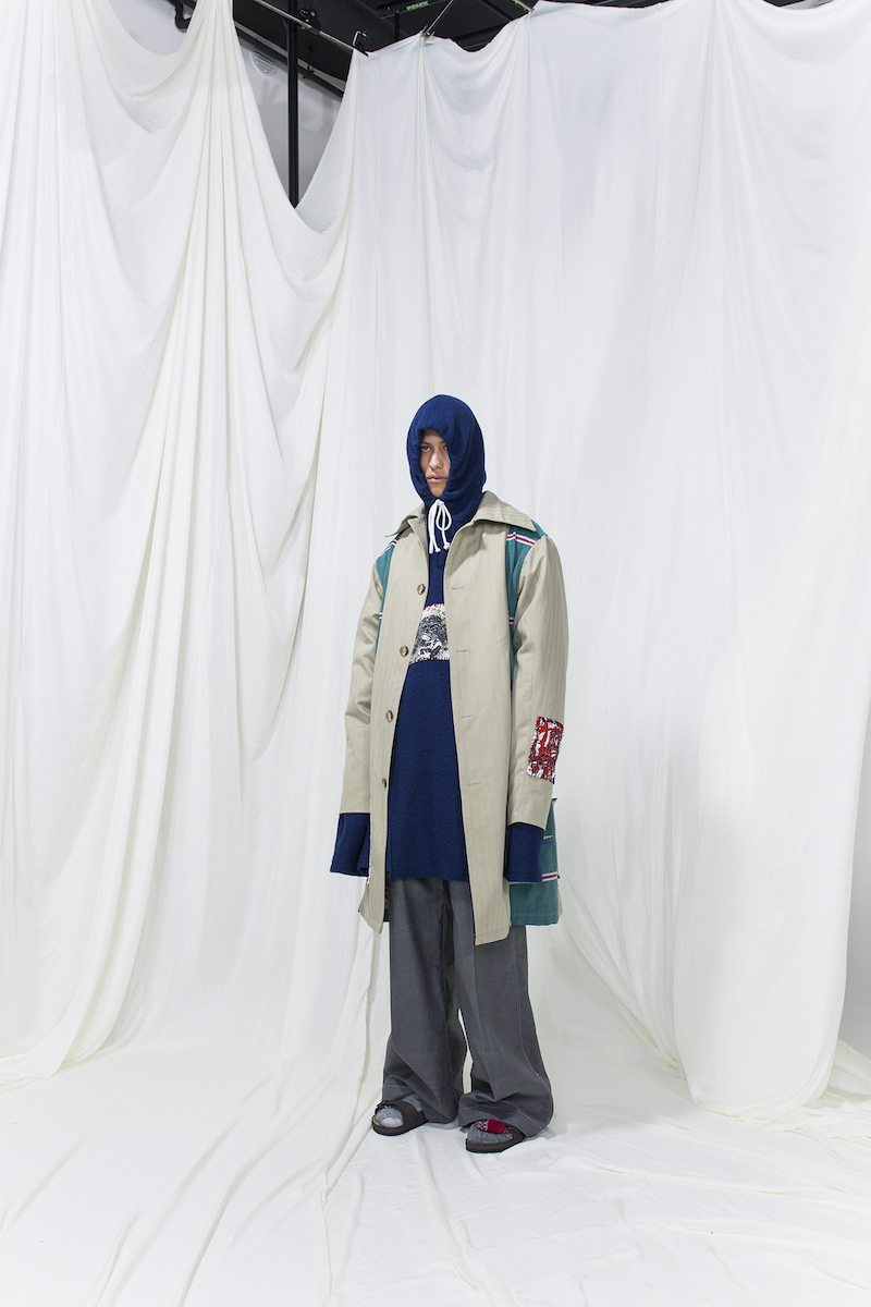 Model is wearing a panelled beige-green coat. Underneath navy blue long hoodie and grey oversized trousers