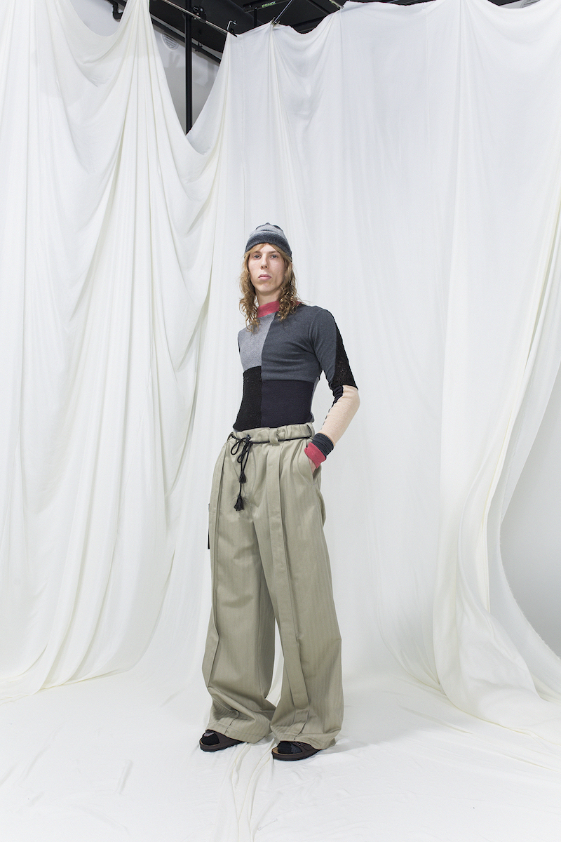 Model is wearing a panelled knitted top with red trimmings, oversized green-beige trousers and grey knitted hat