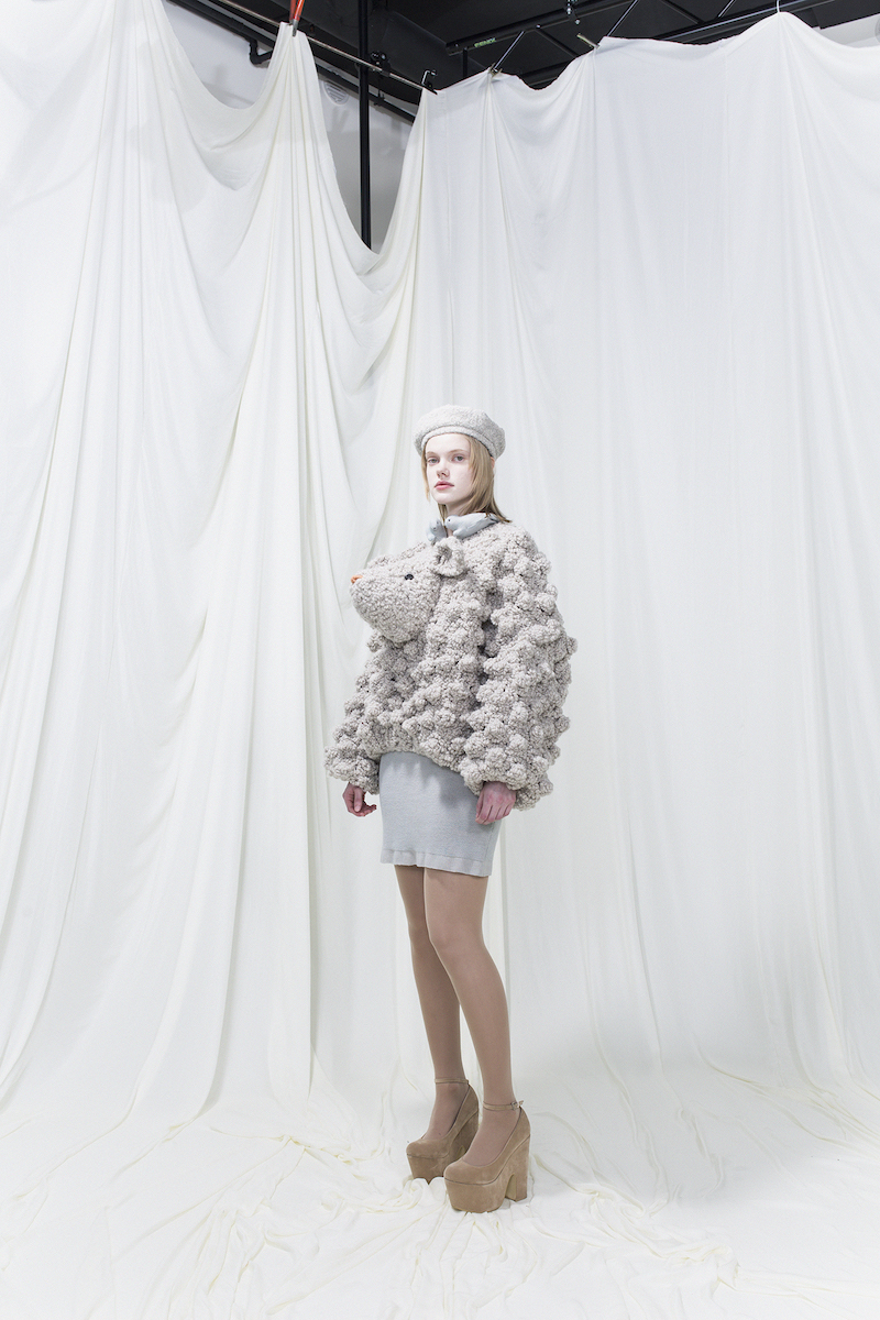Model wearing knitted jumper resembling a sheep with glass eyes and embroidered nose. Grey pique dress underneath. Grey beret.