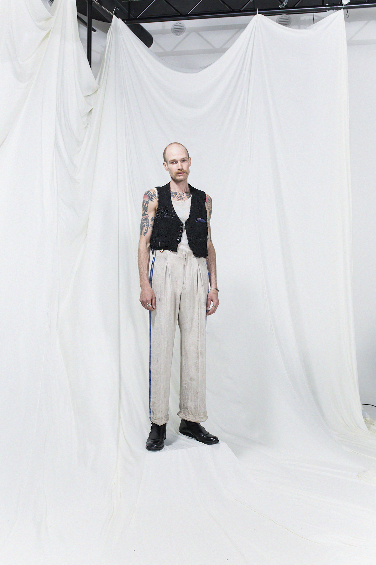 Model is wearing a white tank top, black vest and oversized trousers with blue stripes and suspenders