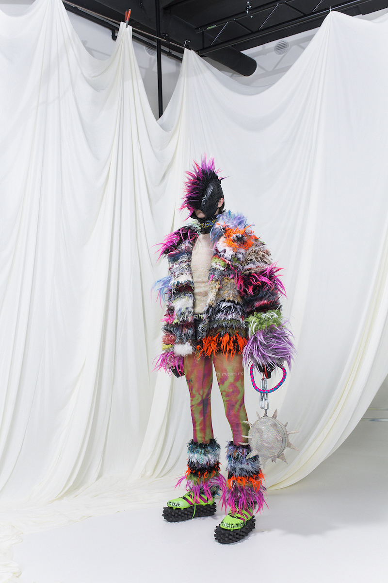 Model is wearing a multicoloured faux-fur coat with matching leg-warmers, multicoloured leggings and a leather choker with a matching mask