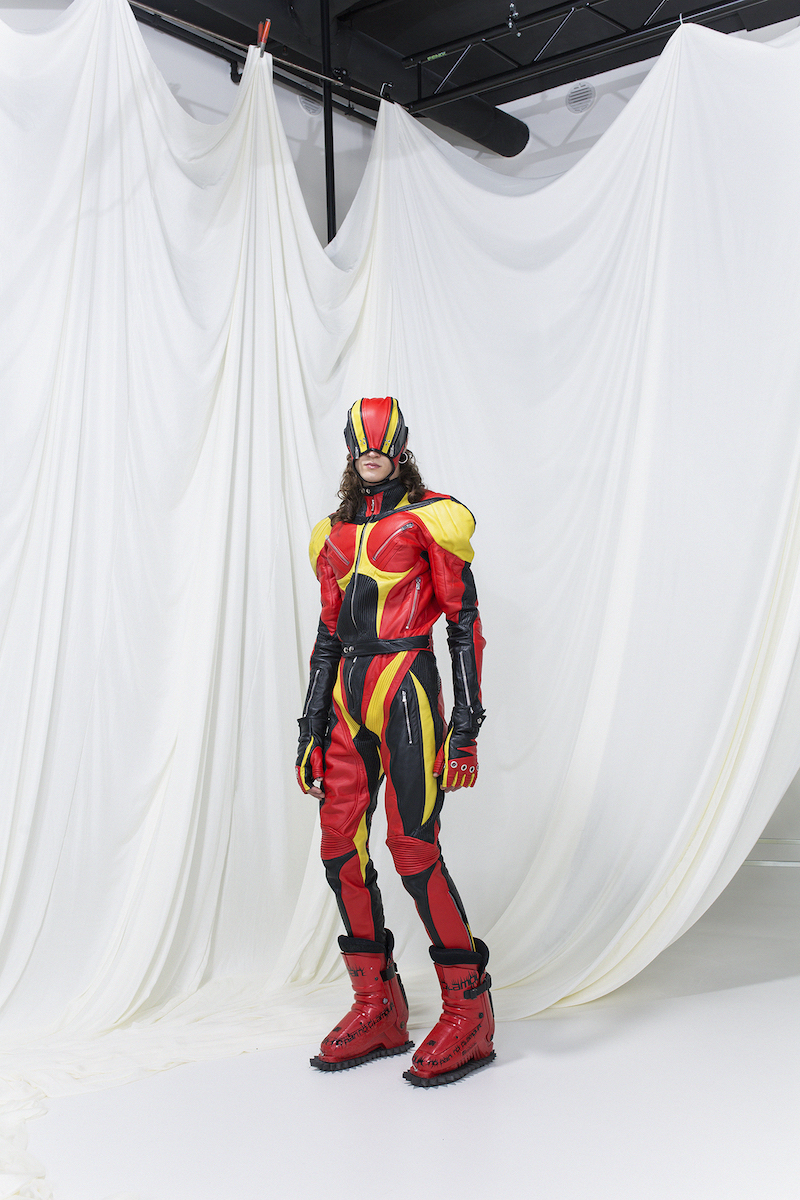 Model is wearing a muscle leather overall in red, yellow and black, with matching gloves and mask. Red boots