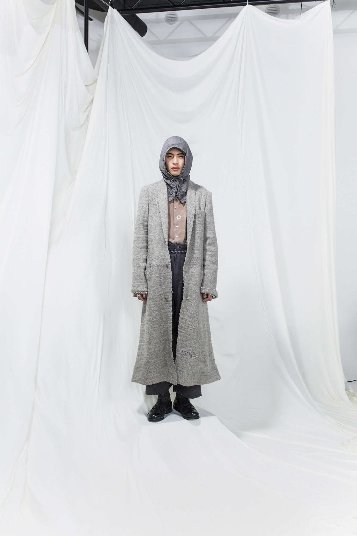 Model is wearing a light grey coat, dark grey hoodie. Underneath pink buttoned shirt and grey oversized trousers