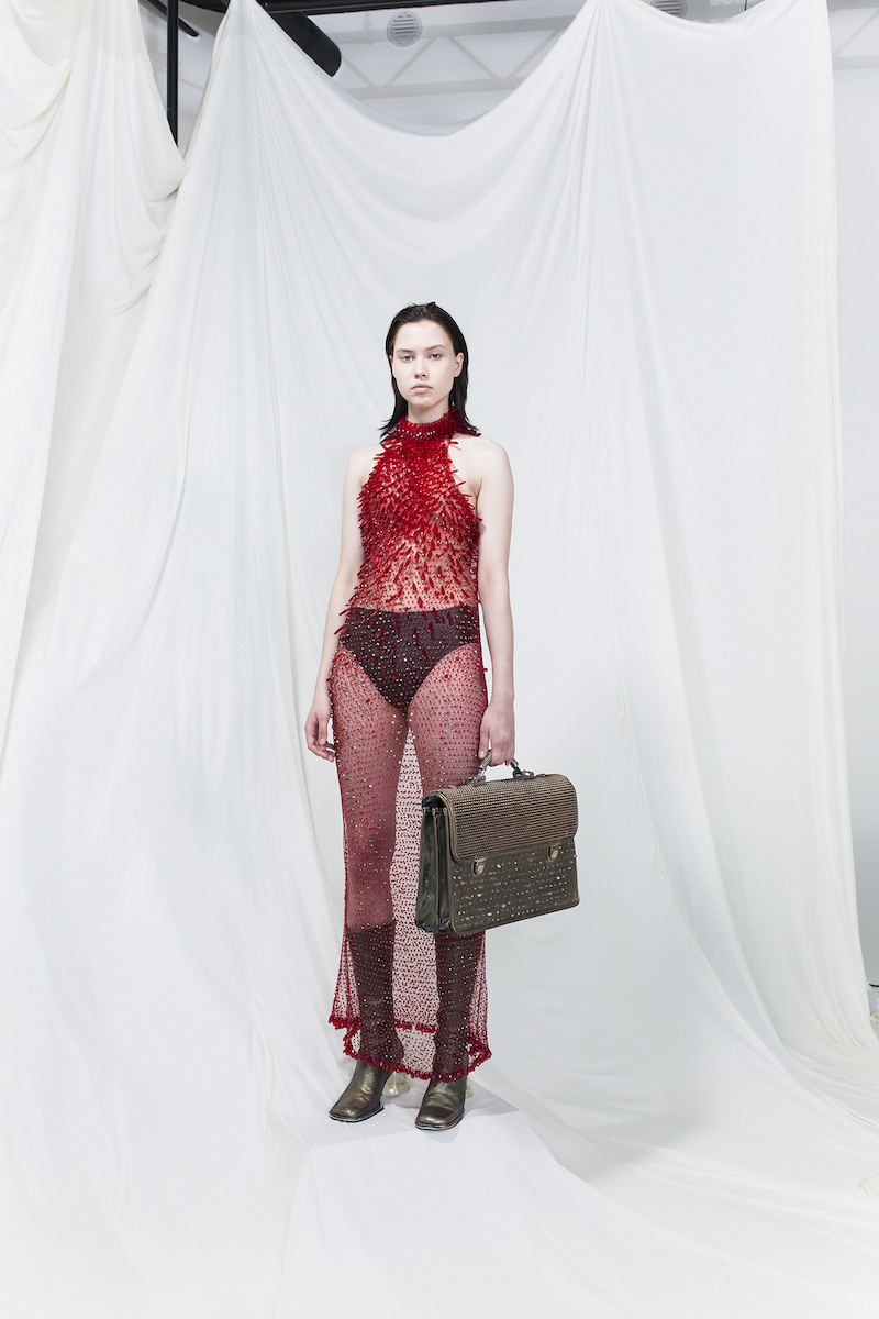 Model is wearing a red sheer long dress with long red plastic crystals. Olive green suitcase and same-coloured heels as accessories