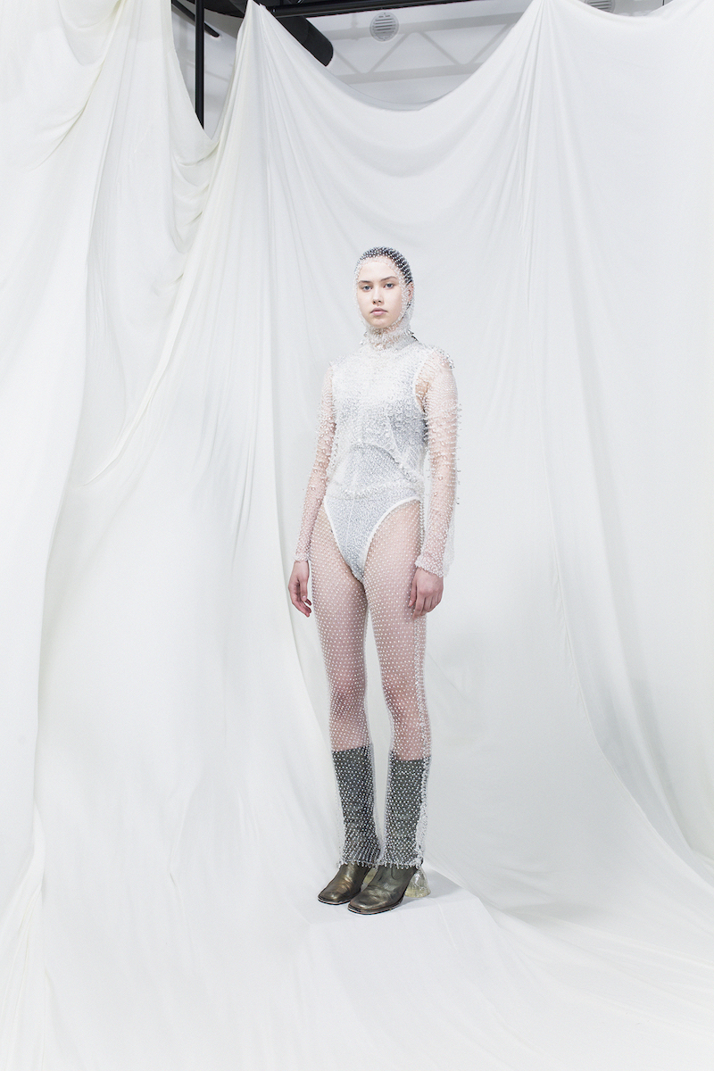 Model is wearing a white bodysuit and sheer spiked dress with olive green heeled boots