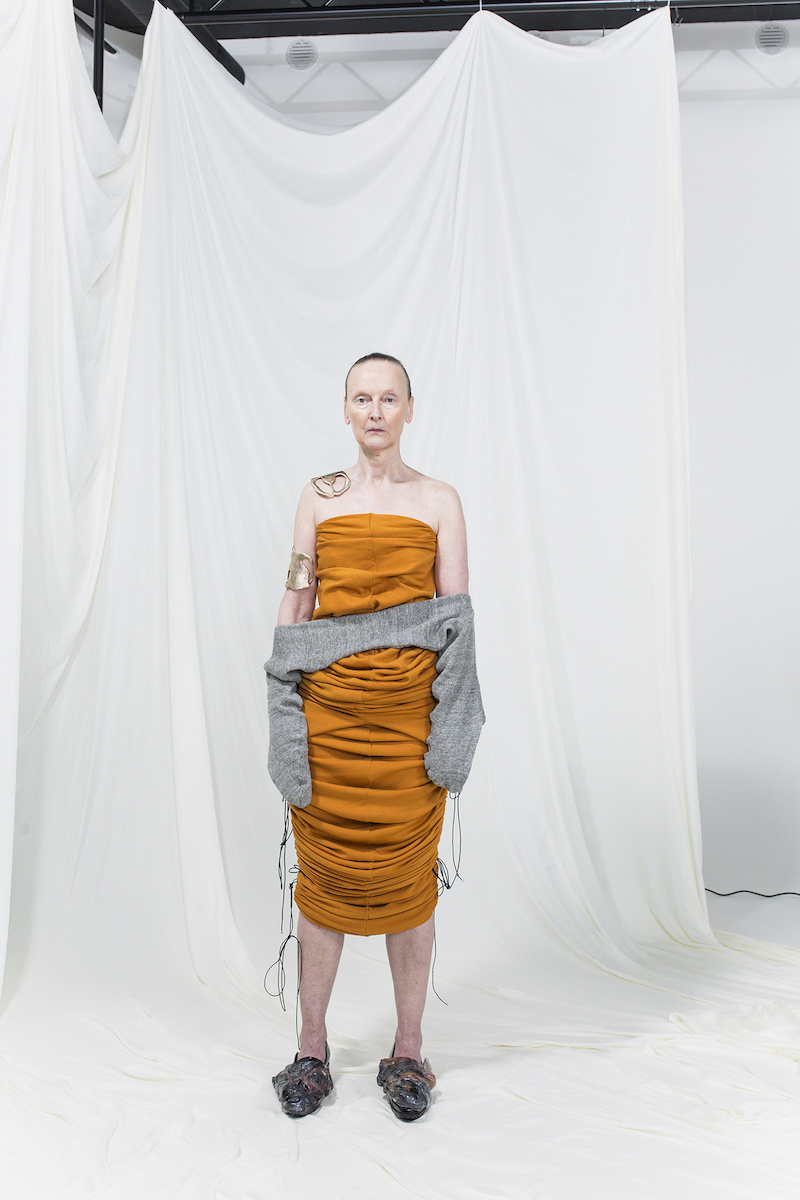 Model is wearing a sleeveless slim draped orange midi-dress with grey infinity-gloves