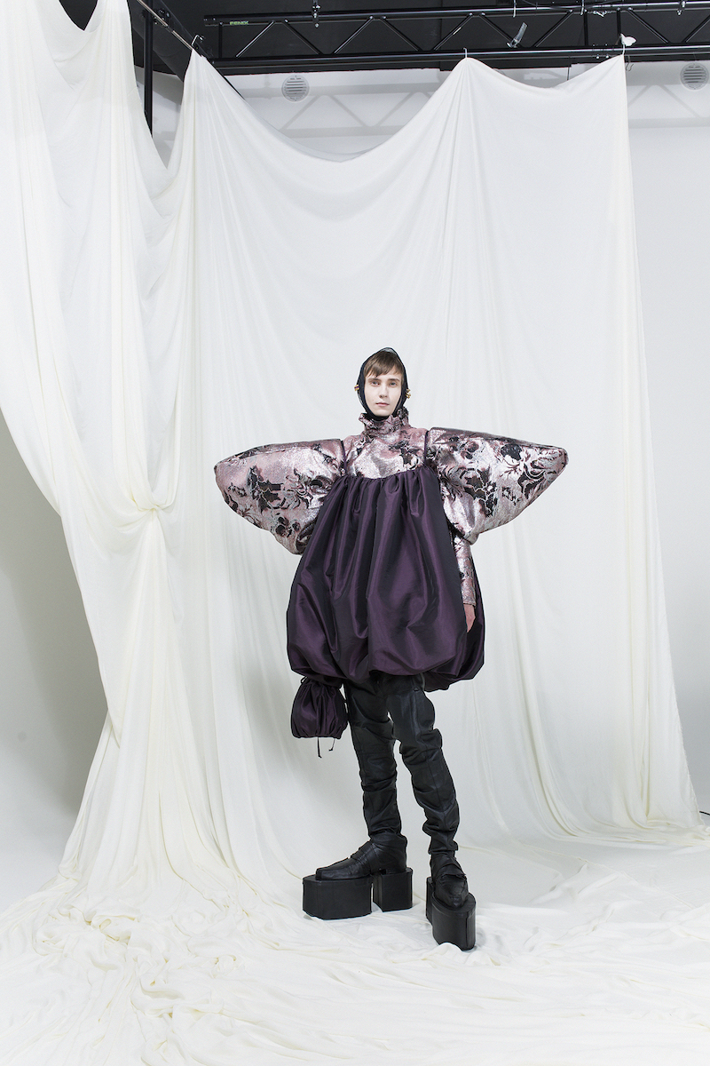 Model is wearing a purple balloon-shaped dress and printed shirt with exaggerated sleeves underneath. Headscarf and long black boots as accessories