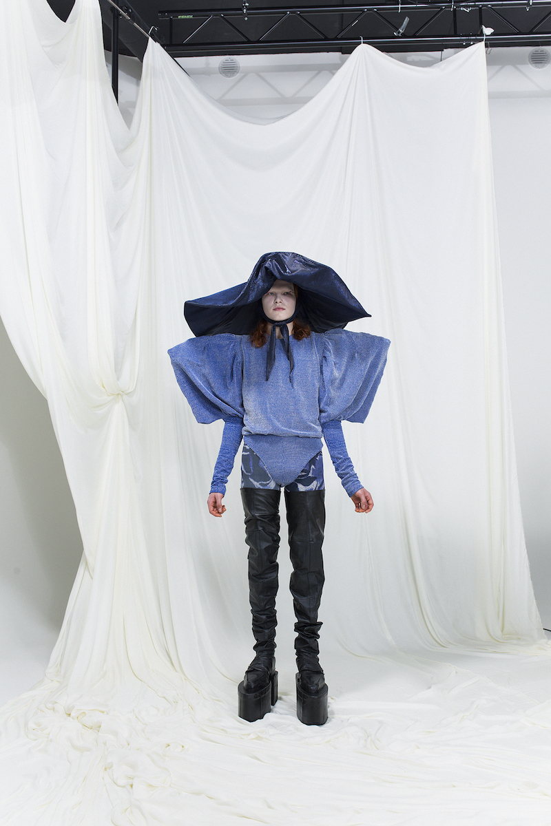 Model is wearing a blue bodysuit with exaggerated shoulders and sleeves. Wide-brim dark blue hat as an accessory