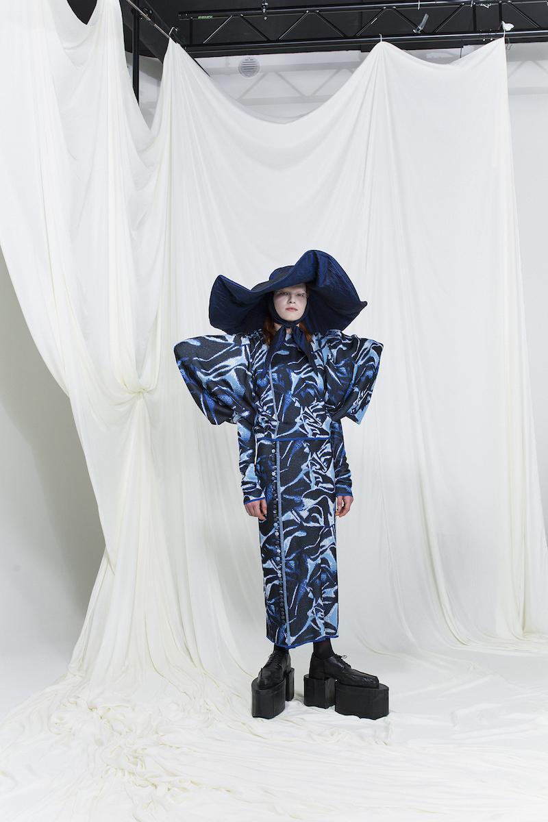 Model is wearing a knitted robe dress with distorted blue pattern and exaggerated shoulders and sleeves. Wide-brim dark blue hat as an accessory