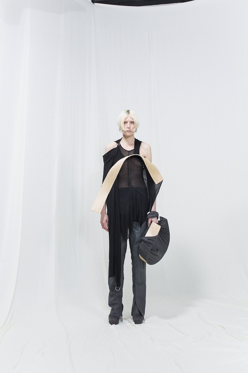 Model is wearing an asymmetrical black top with bended birch piece attached, matching bag and grey trousers underneath