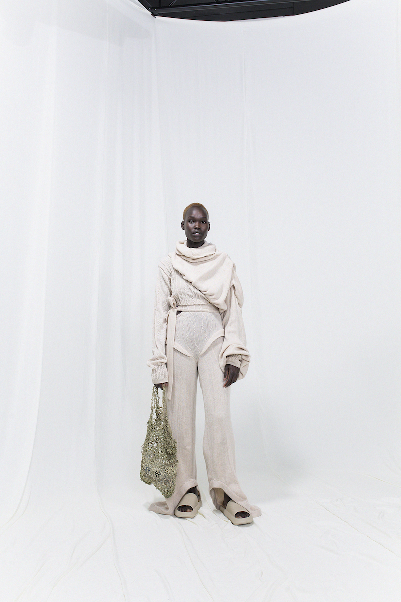 Model is wearing a beige draped and knitted sweater with matching knitted trousers. Slippers and knitted seaweed bag as accessories