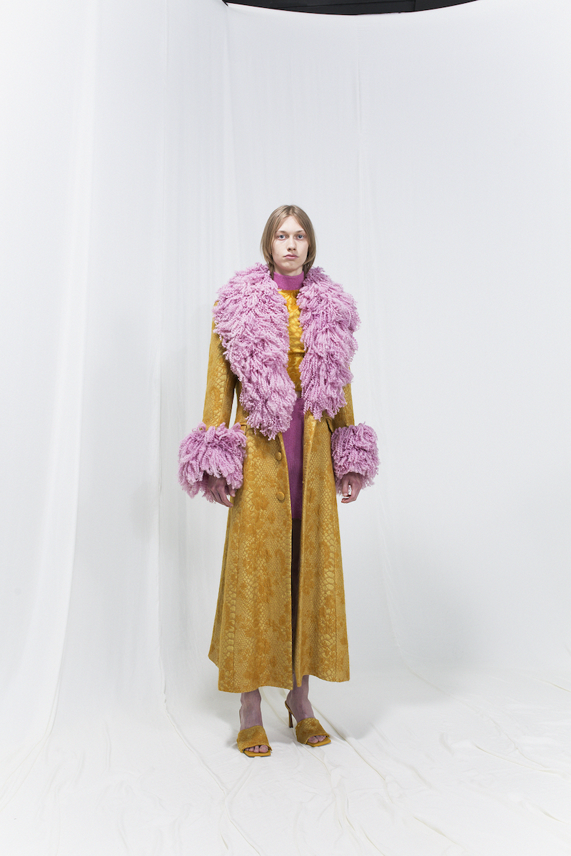 Model is wearing a long ochre coat with flamboyant furry collar and sleeve cuff details