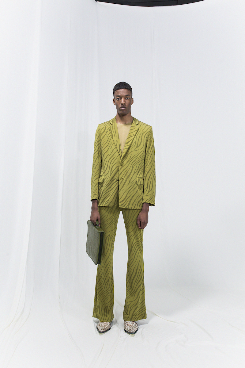 Model wearing an olive green zebra printed jersey blazer, matching bell-bottomed trousers and green suitcase