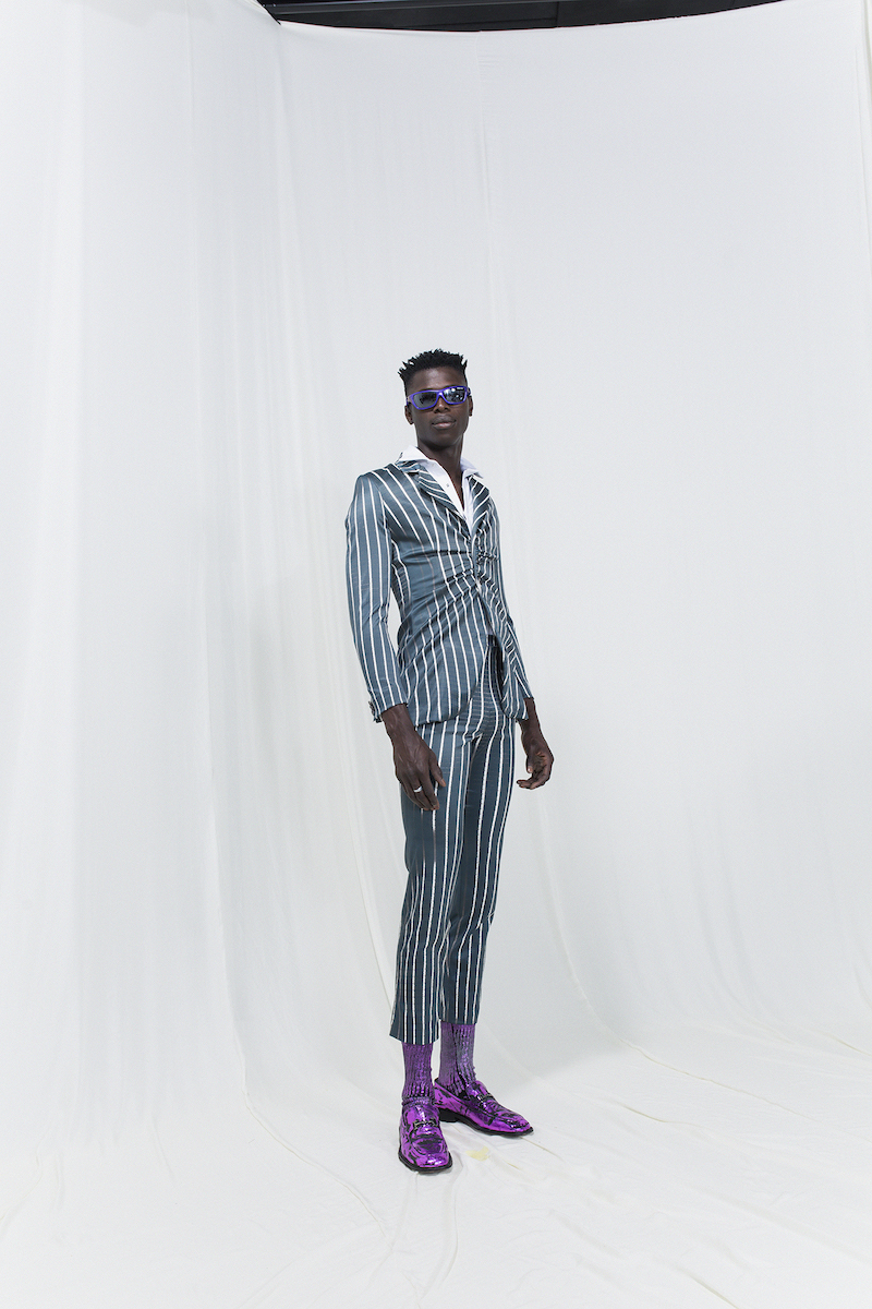 Model is wearing a green striped suit with gatherings and matching cropped trousers. Lilac socks, shoes and sunglasses as accessories